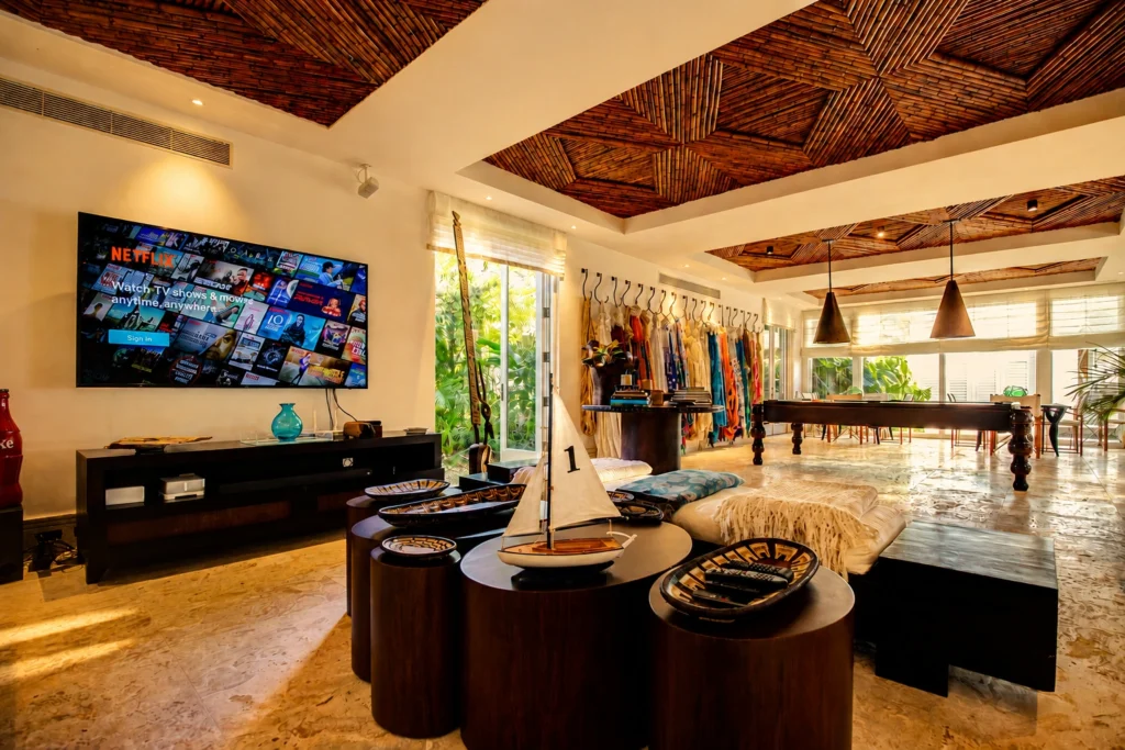 Living room with a wall-mounted Netflix screen showing 'Watch TV shows & movies anytime, anywhere,' a sailboat model on round coffee tables, and a clothing rack along a sunny wall.