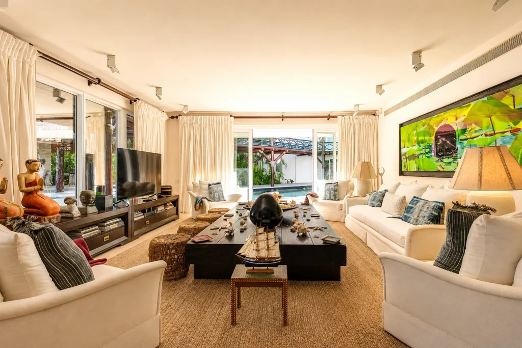Bright living room with white sofas, tropical artwork, and a large black coffee table; sliding glass doors to an outdoor pool area.