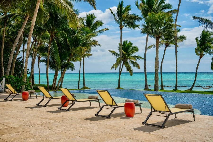 Row of yellow lounge chairs along a sunny poolside with a turquoise sea and palm trees beyond.