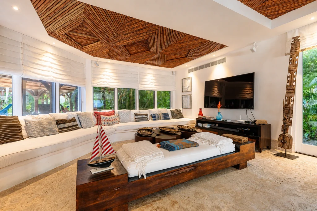 Spacious tropical living room with a white curved sofa, wooden coffee table, and large windows framed by white blinds under a geometric wooden ceiling.