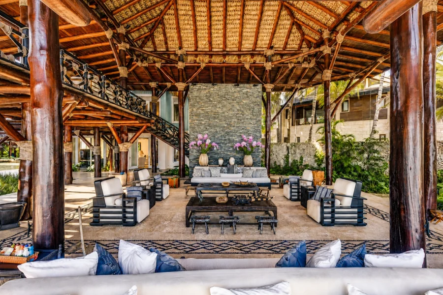 Open-air tropical lounge with dark wooden beams, stone fireplace, and multiple seating areas arranged around a central coffee table.