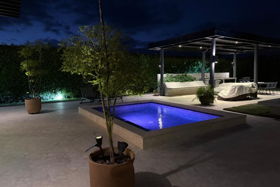 Night-time backyard with a lit blue pool, concrete patio, and a covered outdoor seating area under a pergola.
