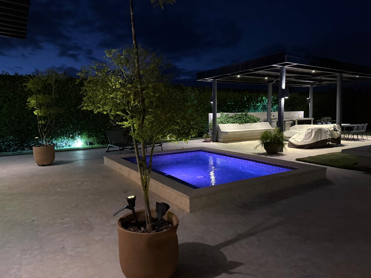 Night-time backyard with a lit blue pool, concrete patio, and a covered outdoor seating area under a pergola.