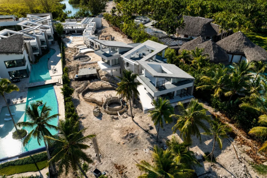 Aerial view of a tropical resort under construction with modern white villas, pools, and palm trees nearby.