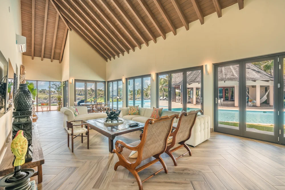 Spacious tropical living room with a wood-beamed vaulted ceiling, wicker chairs, white sofa, and glass doors opening to the pool area.