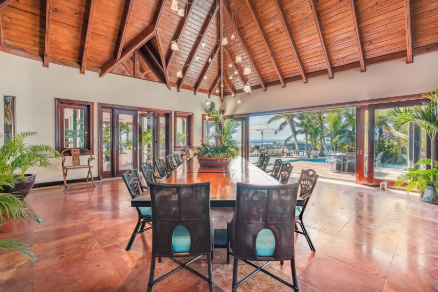 Indoor tropical dining room with a long wooden table, black chairs, and potted plants, opening to a pool and ocean view through glass doors.