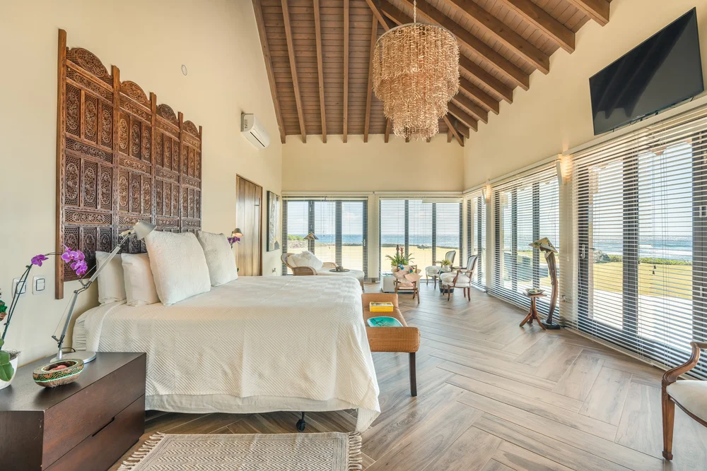 Ocean-view bedroom with a large white bed, carved headboard, and a seating area along glass walls overlooking the beach.